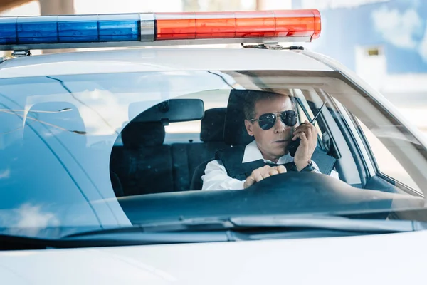 Serious middle aged policeman in sunglasses talking on radio set in car — Stock Photo