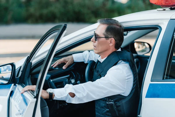Visão lateral do policial de meia idade do sexo masculino em colete à prova de balas e óculos de sol saindo do carro — Fotografia de Stock