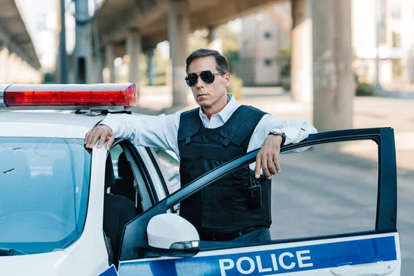 Confident mature policeman in sunglasses and bulletproof vest standing near car at city street — Stock Photo