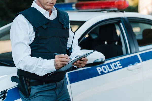 Vista parcial del oficial de policía masculino de mediana edad con un chaleco antibalas escrito en portapapeles cerca del coche en la calle - foto de stock