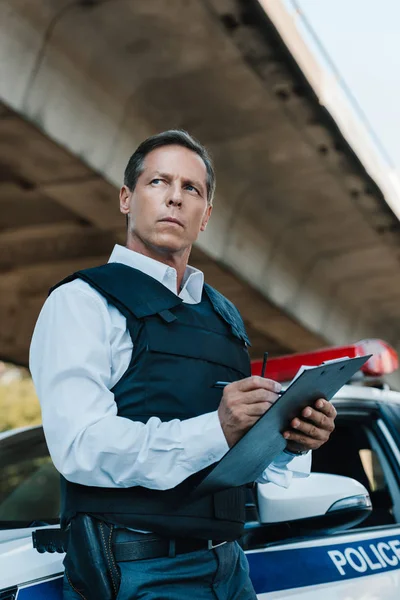 Low angle view of middle aged male police officer in bulletproof vest writing in clipboard near car at street — Stock Photo
