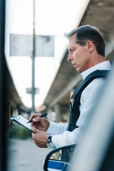 Side view of middle aged male police officer in bulletproof vest writing in clipboard near car at street — Stock Photo