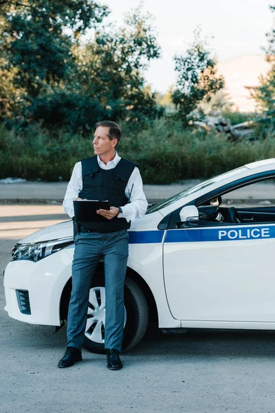 Mature male police officer in bulletproof vest writing in clipboard near car at street — Stock Photo