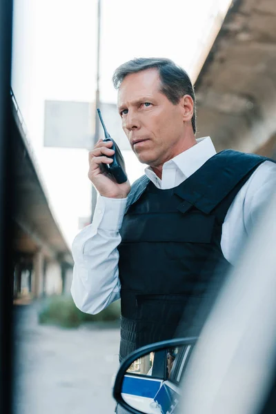 Mature male police officer in bulletproof vest talking on radio set near car at street — Stock Photo