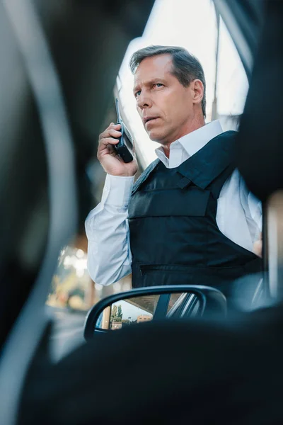 Low angle view of serious mature male police officer in bulletproof vest talking on radio set near car at street — Stock Photo