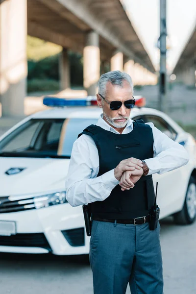 Männlicher Polizeibeamter mittleren Alters in Sonnenbrille und schusssicherer Weste bei der Kontrolle der Armbanduhr an der Stadtstraße — Stockfoto