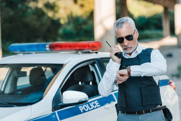 Middle aged policeman in bulletproof vest and sunglasses checking wristwatch near car at street — Stock Photo