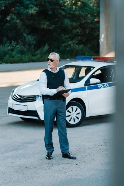 Mature policeman in sunglasses and bulletproof vest writing in clipboard near car at street — Stock Photo