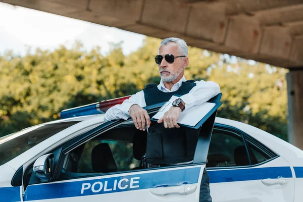 Oficial de policía masculino en chaleco antibalas y gafas de sol de pie con portapapeles cerca del coche en la calle - foto de stock