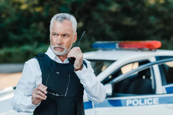 Serious middle aged policeman in bulletproof vest talking on radio set near car at street — Stock Photo