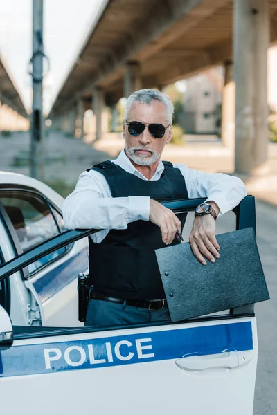 Serious mature policeman in bulletproof vest and sunglasses standing with clipboard near car at street — Stock Photo
