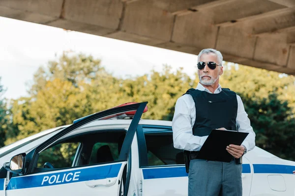 Policier en gilet pare-balles et lunettes de soleil écrit dans le presse-papiers près de la voiture à la rue — Photo de stock