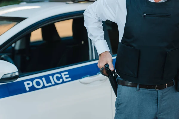 Cropped image of policeman in bulletproof vest taking off gun from holster near car at street — Stock Photo