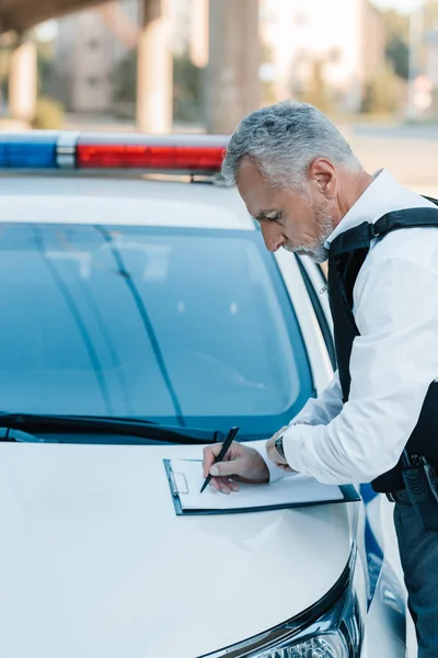 Vista lateral del oficial de policía masculino enfocado escribiendo en portapapeles en el coche en la calle - foto de stock