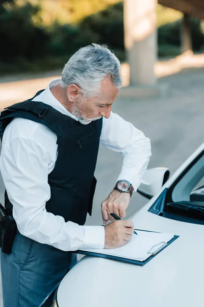 Middle aged policeman looking at wristwatch and writing in clipboard on car at street — Stock Photo