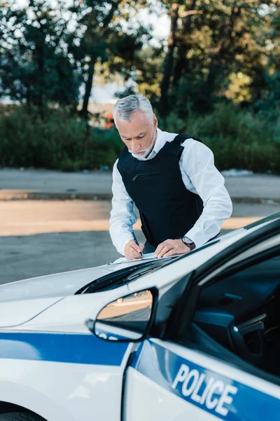 Concentrated mature male police officer writing in clipboard on car at street — Stock Photo