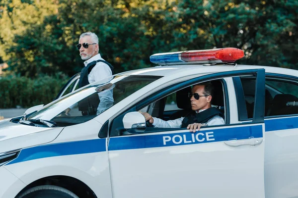 Middle aged policemen in sunglasses walking out from car at urban street — Stock Photo