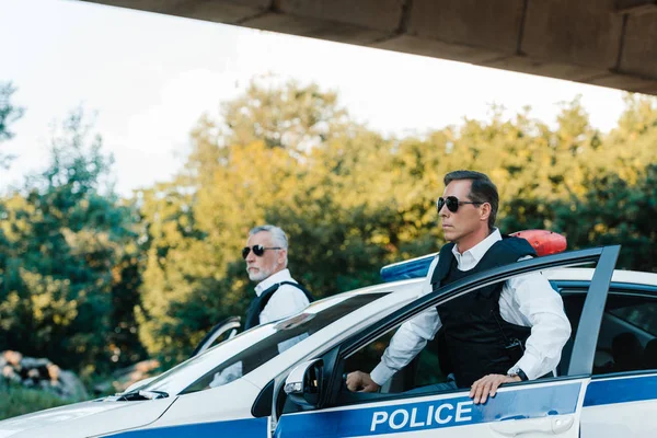 Enfoque selectivo de policía maduro en gafas de sol y chalecos antibalas de pie cerca de coche en la calle de la ciudad - foto de stock