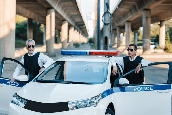 Confident mature policeman in sunglasses and bulletproof vests standing near car at city street — Stock Photo