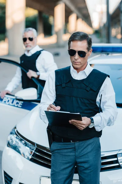 Male police officer in sunglasses writing in clipboard while his colleague standing near car at street — Stock Photo