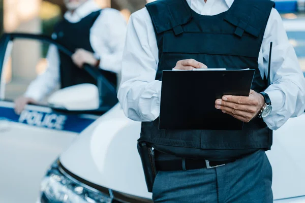 Abgeschnittenes Bild eines männlichen Polizisten, der in Klemmbrett schreibt, während sein Kollege in der Nähe von Auto auf der Straße steht — Stockfoto