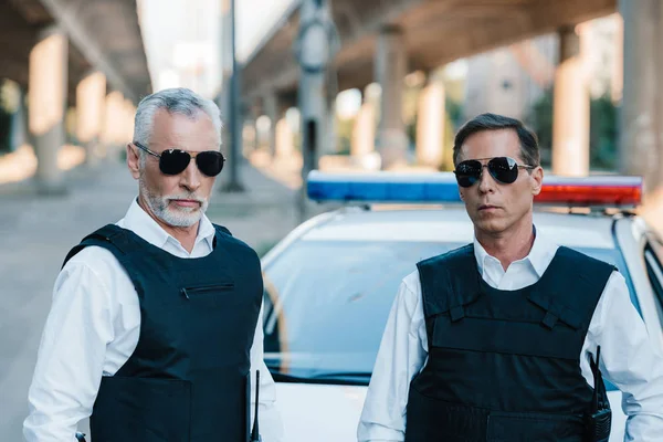 Portrait of serious police officers in sunglasses and bulletproof vests near car at street — Stock Photo