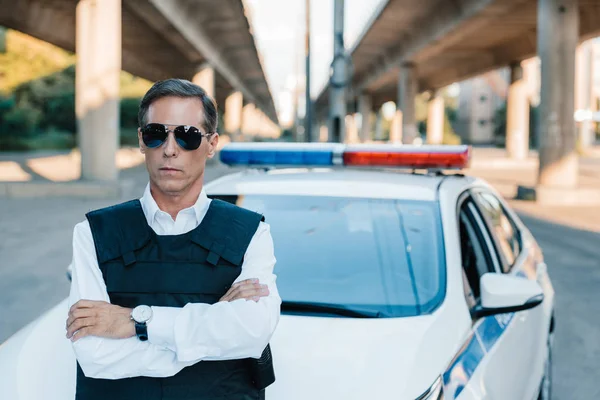 Mature policeman in sunglasses and bulletproof vest standing with crossed arms near car at street — Stock Photo