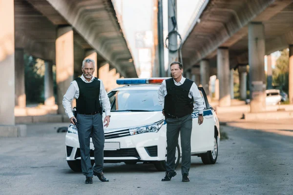 Schwerer Polizist zieht Waffe aus Holster, während sein Kollege neben Auto auf der Straße steht — Stockfoto