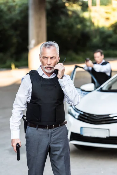 Mature policeman with handgun talking on radio set while his colleague aiming by gun behind at street — Stock Photo