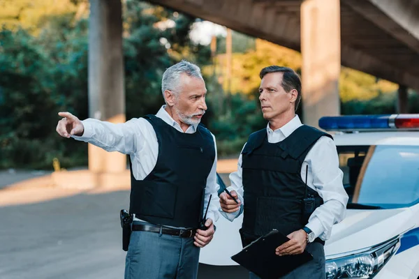 Mature male police officer pointing by finger to colleague with clipboard near car at street — Stock Photo