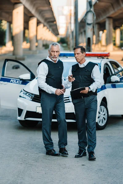 Policier masculin avec presse-papiers pointant du doigt un collègue debout près de la voiture dans la rue — Photo de stock