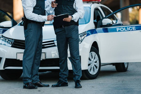 Partial view of male police officer with clipboard and pointing by finger to colleague standing near car at street — Stock Photo