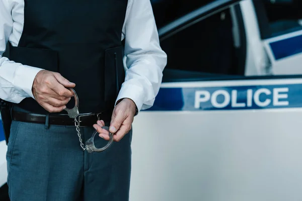 Частичный вид полицейского мужчины, держащего наручники возле машины на улице — стоковое фото