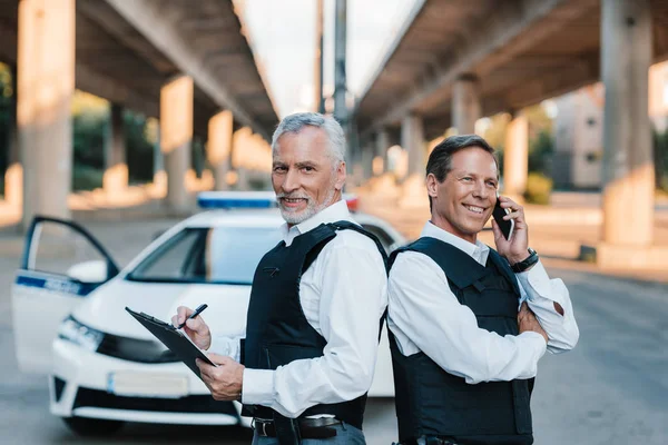 Sorridente ufficiale di polizia maschile che parla su smartphone mentre il suo collega scrive negli appunti in auto in strada — Foto stock