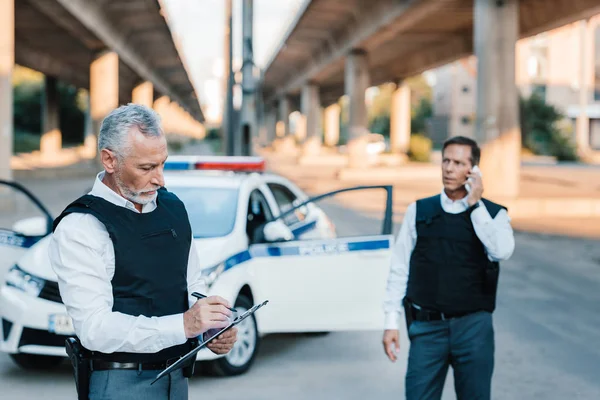 Middle aged police officer writing in clipboard while his colleague talking on smartphone at street — Stock Photo