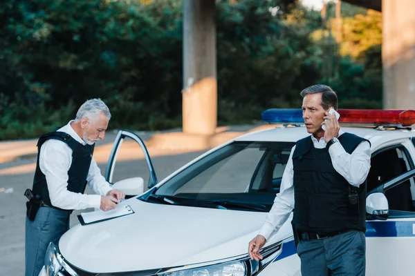 Mature police officer talking on smartphone while his colleague writing in clipboard on car at street — Stock Photo