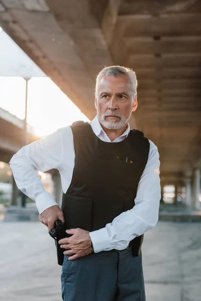 Middle aged male police officer in bulletproof vest taking off gun from holster at city street — Stock Photo