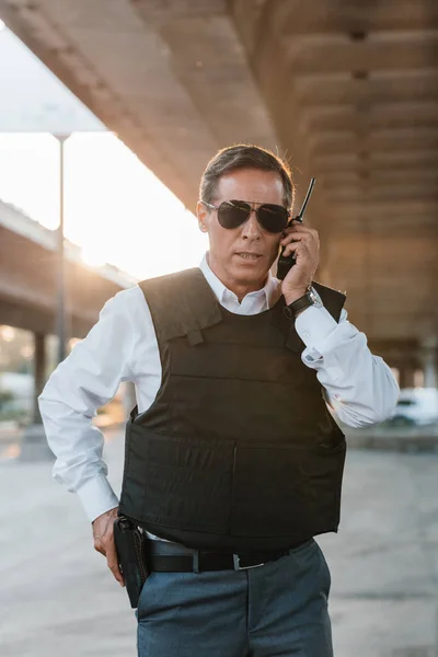 Oficial de policía masculino en gafas de sol hablando en el set de radio y quitando la pistola de la funda en la calle de la ciudad - foto de stock