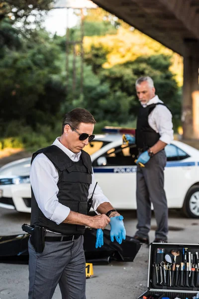 Maduro policía en gafas de sol ponerse guantes de látex mientras su colega de pie detrás de la escena del crimen con el cadáver en bolsa de cuerpo - foto de stock