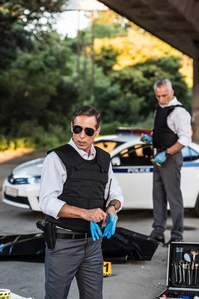 Policía serio en gafas de sol ponerse guantes de látex mientras su colega de pie detrás de la escena del crimen con el cadáver en la bolsa del cuerpo - foto de stock