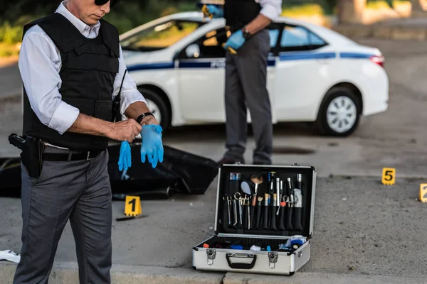 Visão parcial de policial do sexo masculino em óculos de sol colocando luvas de látex, enquanto seu colega de pé atrás na cena do crime com cadáver no saco do corpo — Fotografia de Stock