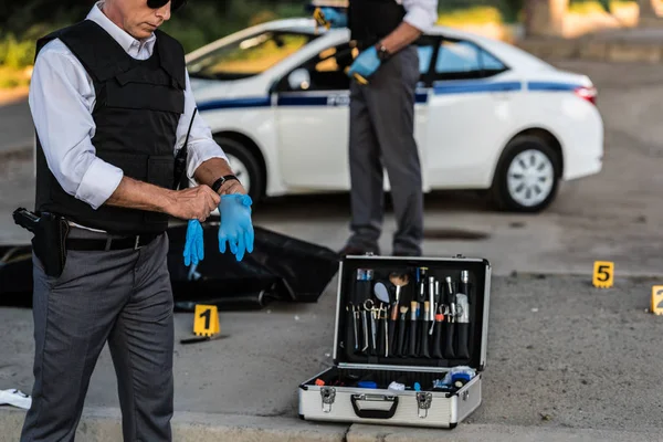 Vista parcial del oficial de policía masculino en gafas de sol que se pone guantes de látex mientras su colega está parado detrás de la escena del crimen con el cadáver en la bolsa del cuerpo - foto de stock