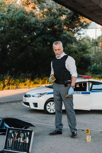 Oficial de policía masculino concentrado en guantes de látex en la escena del crimen con el cadáver en la bolsa del cuerpo - foto de stock