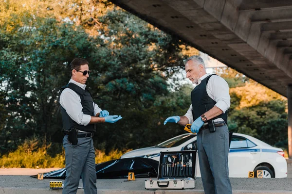 Side view of policemen in latex gloves at crime scene with case for investigation tools and corpse in body bag — Stock Photo