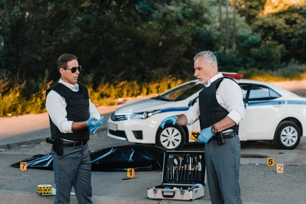 Male police officer in latex gloves pointing by finger to colleague in sunglasses standing near at crime scene with corpse in body bag — Stock Photo