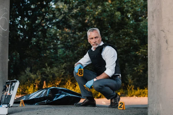 Oficial de policía masculino serio en guantes de látex sentado en la escena del crimen con el cadáver en la bolsa del cuerpo - foto de stock