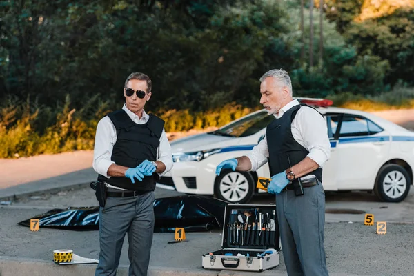 Mature policeman in latex gloves pointing by finger to colleague in sunglasses standing near at crime scene with corpse in body bag — Stock Photo