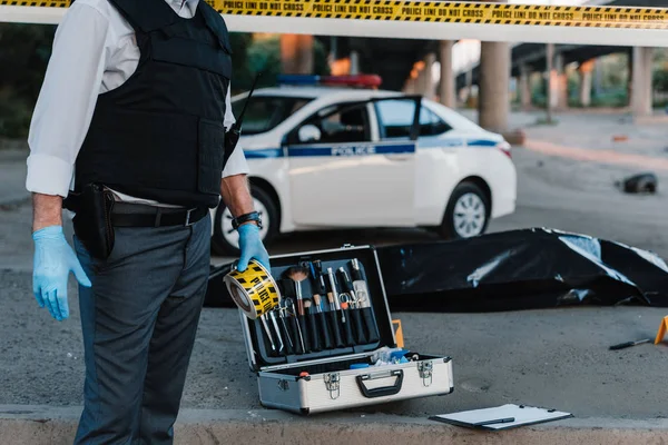 Частичный вид полицейского в латексных перчатках, держащего полицейскую линию на месте преступления с трупом в мешке для трупов — стоковое фото
