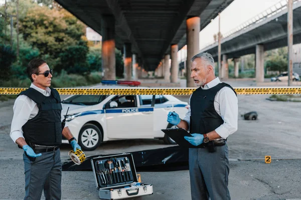 Зрелый полицейский с планшетом разговаривает с коллегой в солнечных очках рядом с трупом в мешке для трупов на месте преступления — стоковое фото