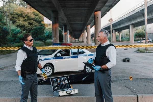 Mature policeman with clipboard talking to colleague in sunglasses near corpse in body bag at crime scene — Stock Photo